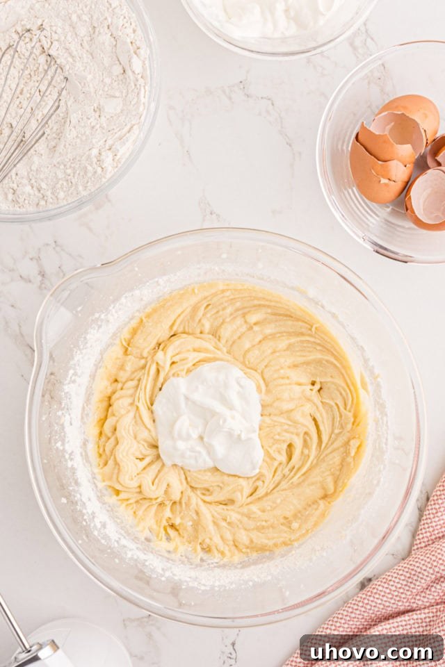A visual showing creamy sour cream being added to the partially mixed strawberry pound cake batter in a glass bowl, emphasizing the alternation with dry ingredients.