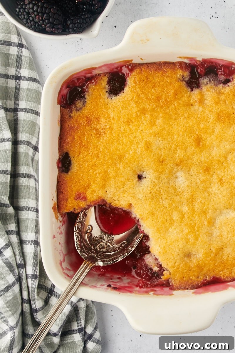 Overhead view of a freshly baked blackberry cobbler in a baking dish, with a serving portion removed and a spoon resting nearby, revealing the bubbly fruit and golden crust.