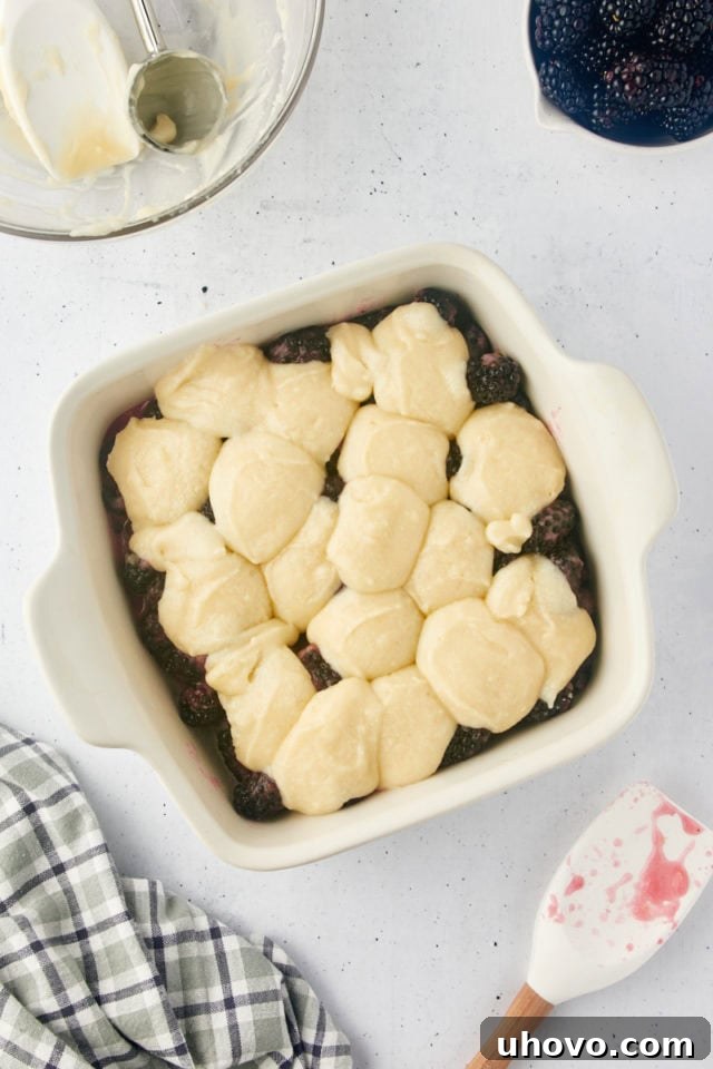 Overhead view of the cobbler topping batter being carefully spooned in dollops over the vibrant blackberry filling in a baking dish, ready for baking to golden perfection.