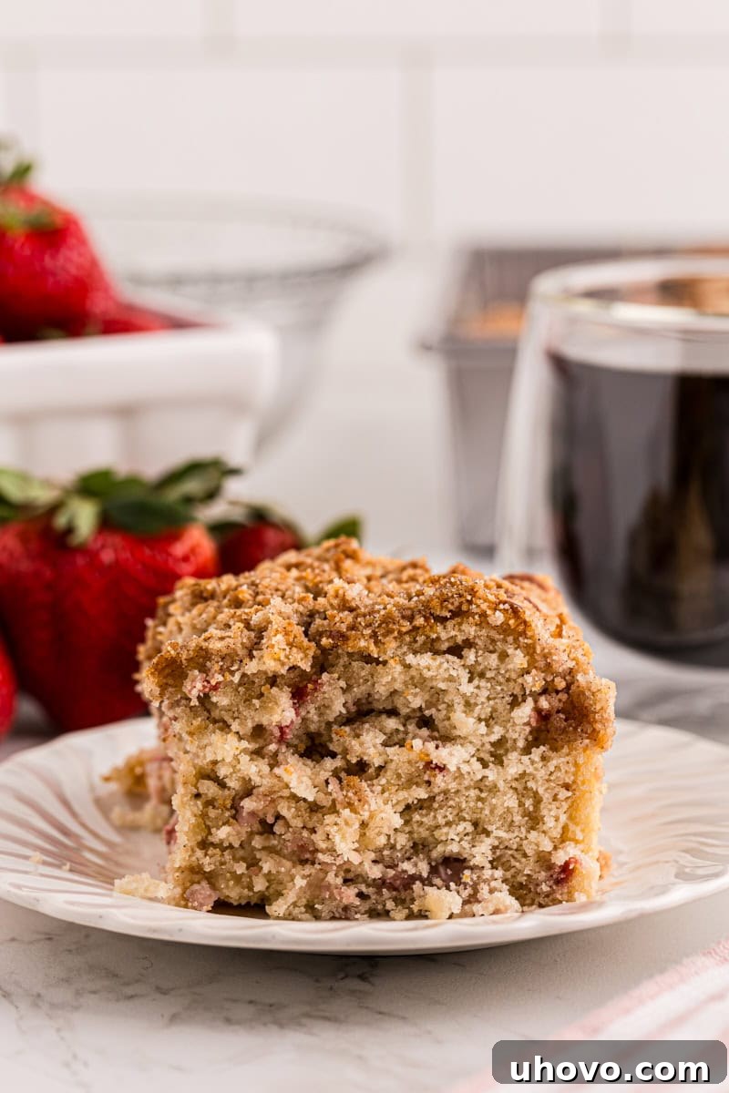 a slice of strawberry coffee cake on a white plate with a cup of coffee and fresh strawberries in the background