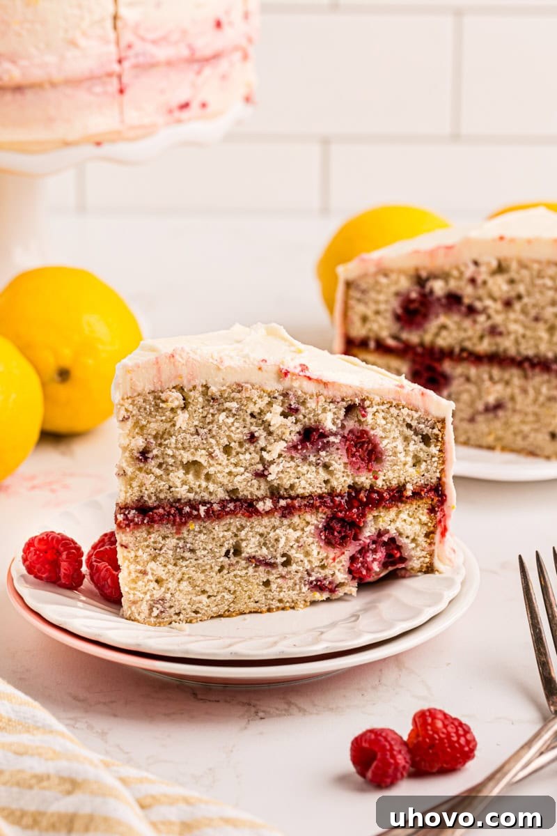 A beautifully decorated slice of lemon raspberry cake on a white plate, with more of the cake in the background.