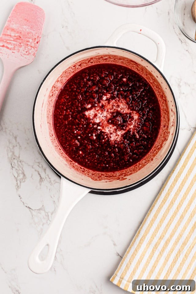 Overhead view of a cornstarch slurry being added to the cooked raspberry mixture in a saucepan.