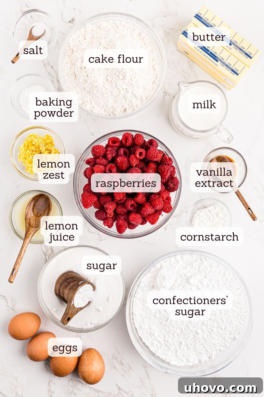 Overhead view of various ingredients for lemon raspberry cake laid out on a surface, including flour, sugar, butter, eggs, lemons, and fresh raspberries.