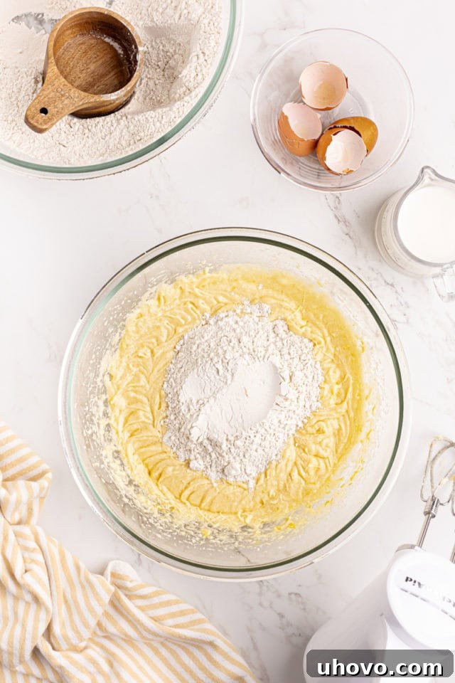 Overhead view of dry ingredients being added in portions to the wet cake batter in a glass mixing bowl.