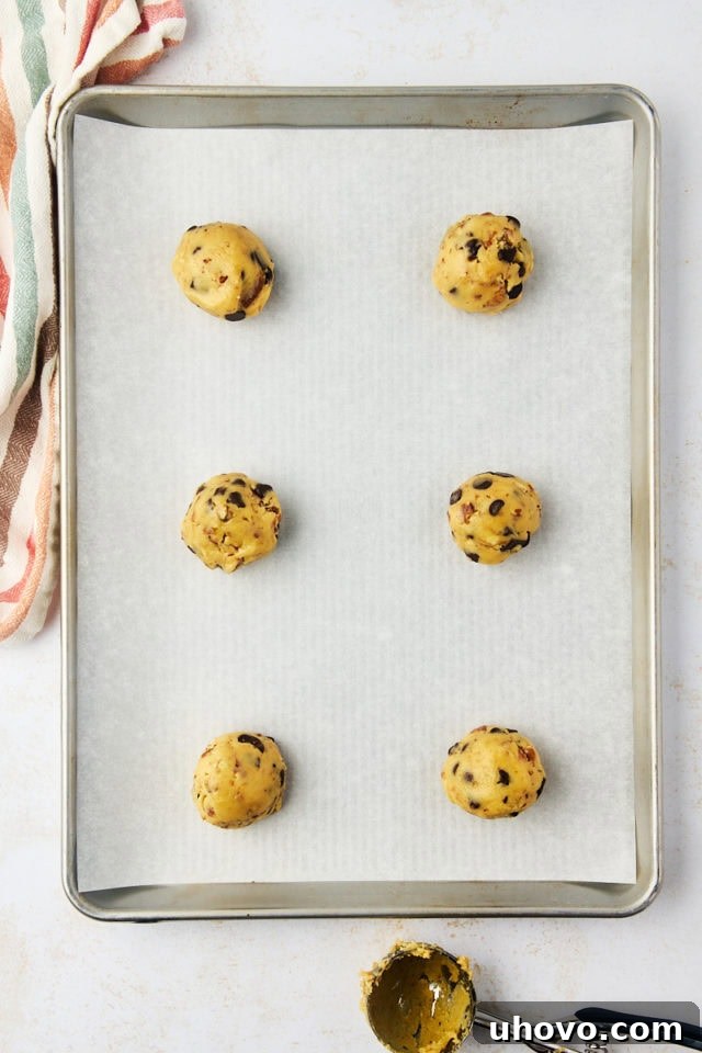 Overhead view of portioned thick chocolate chip cookie dough balls neatly arranged on a parchment-lined baking sheet, ready for a second chill.