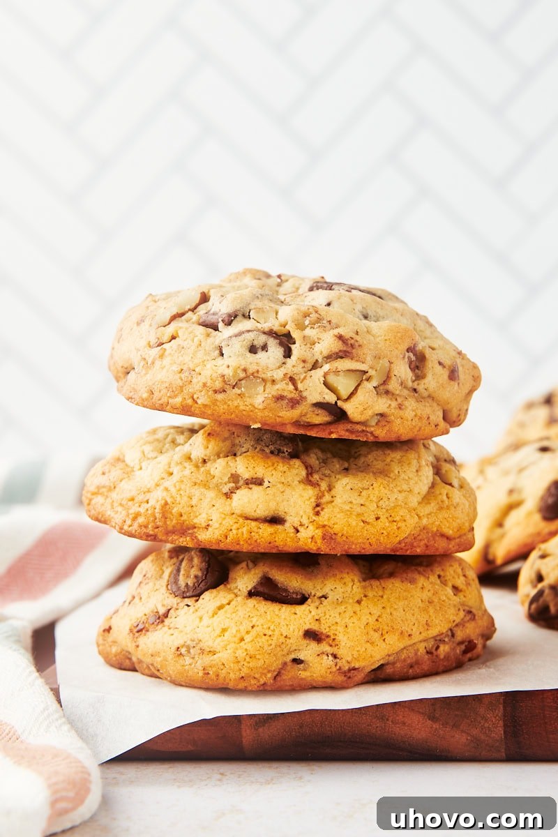 A stack of three thick chocolate chip cookies on a rustic wooden board, highlighting their height and rich appearance.