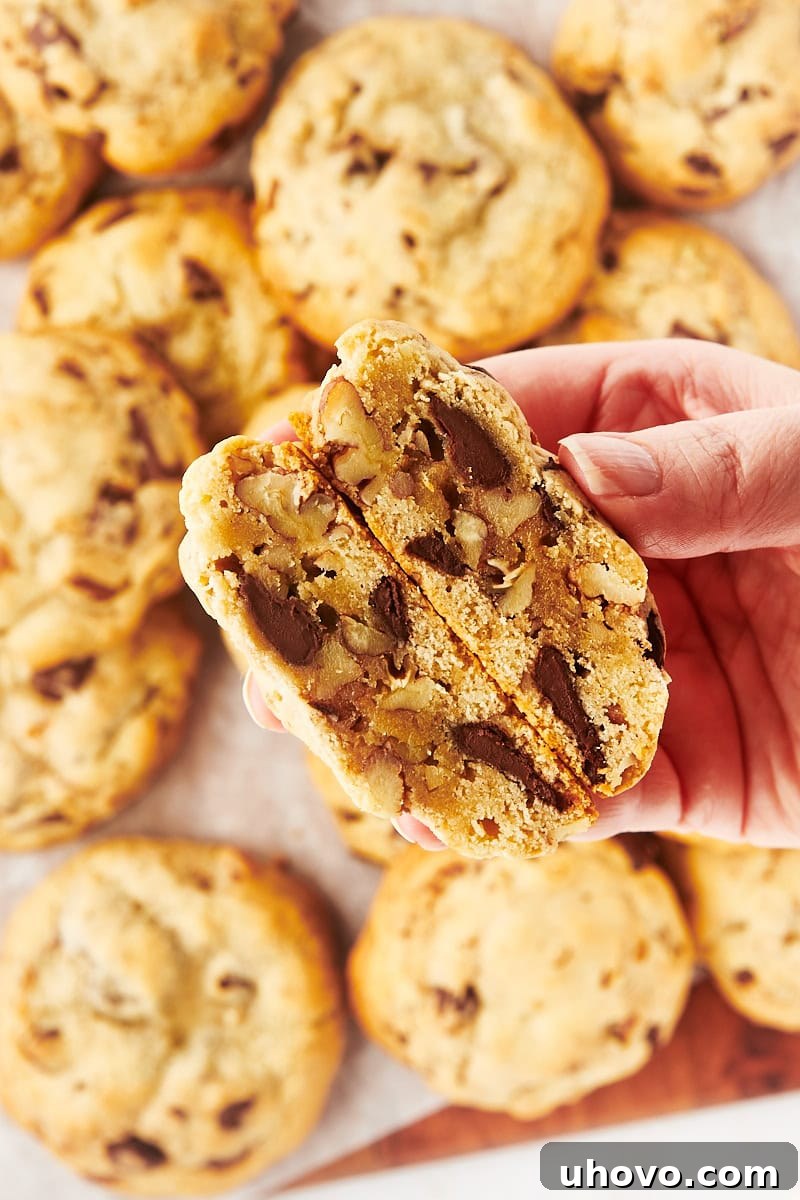A hand holding two halves of a thick chocolate chip cookie, revealing its rich, gooey center, with more cookies blurred in the background.