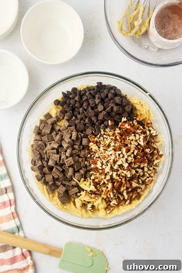 Overhead view of chocolate chips and toasted nuts being folded into cookie dough in a glass bowl.