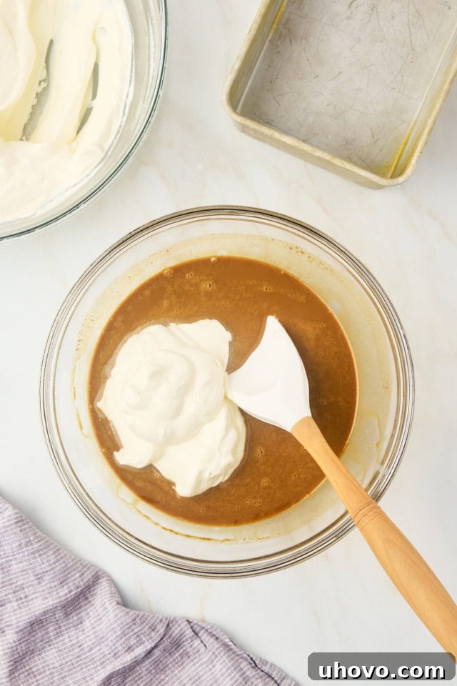 Overhead view of a portion of fluffy whipped cream being gently folded into the rich coffee and condensed milk mixture in a clear glass bowl, illustrating the initial blending step.