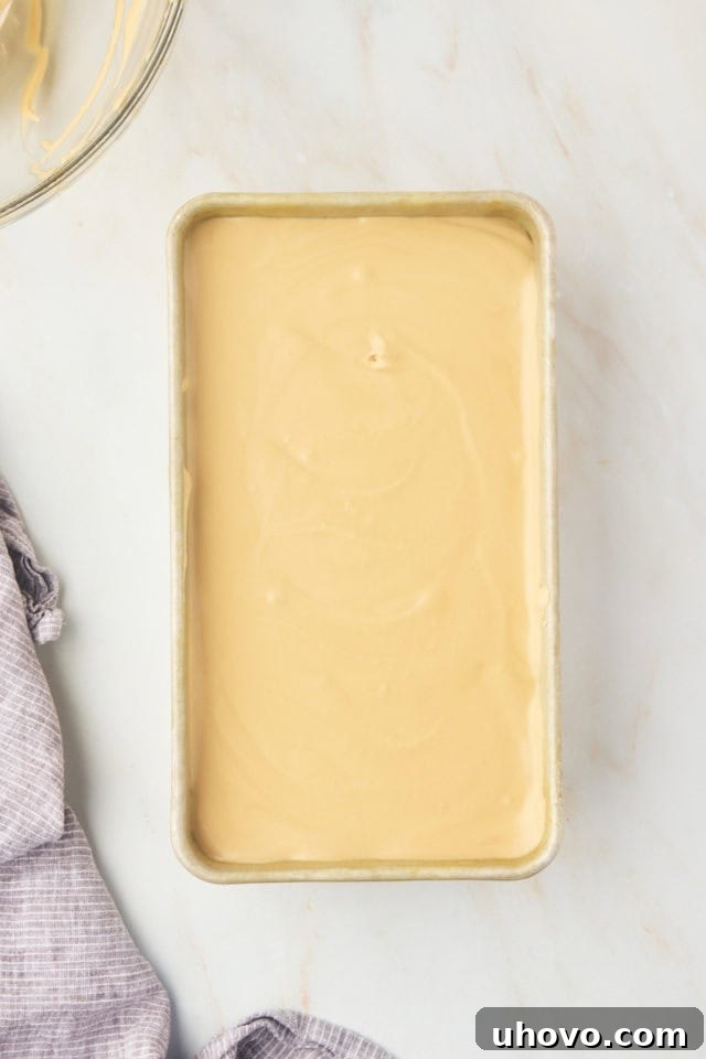 Overhead view of the creamy coffee ice cream mixture smoothed into a cold metal loaf pan, covered with plastic wrap, and ready for freezing.