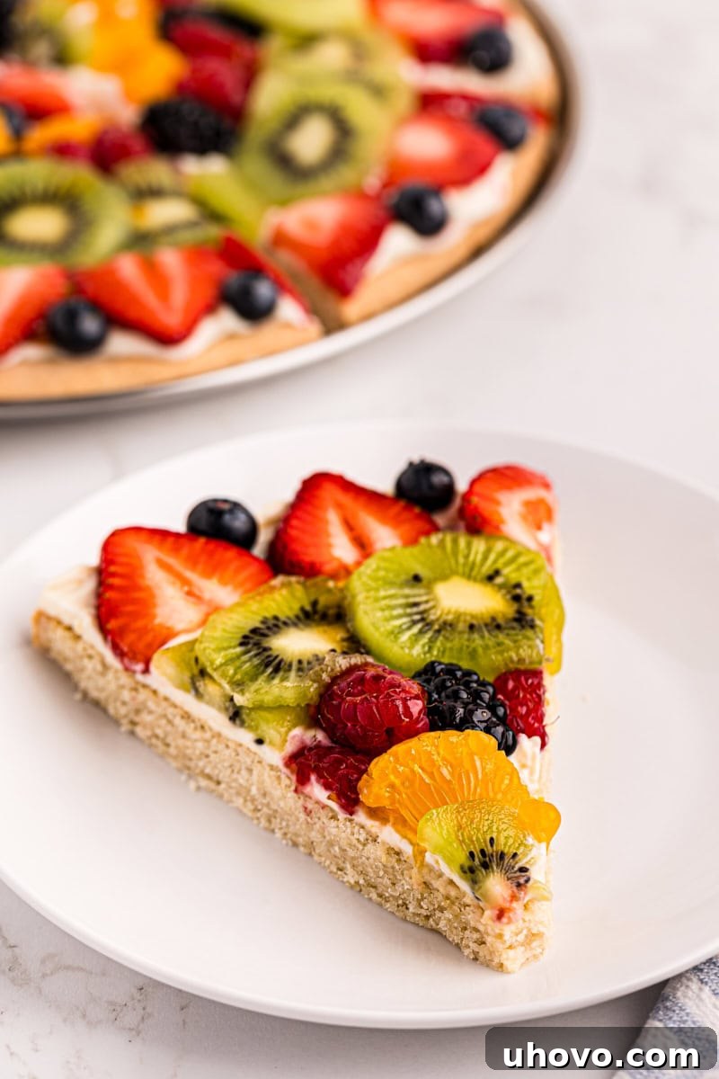 a slice of fruit pizza on a white plate with the remaining pizza in the background