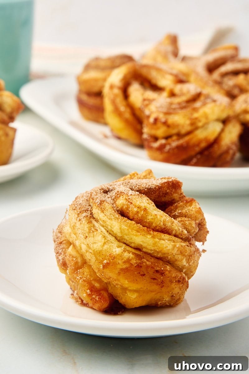 A perfectly golden-brown cruffin on a white plate, with more cruffins in the background, showcasing its flaky layers and inviting texture.