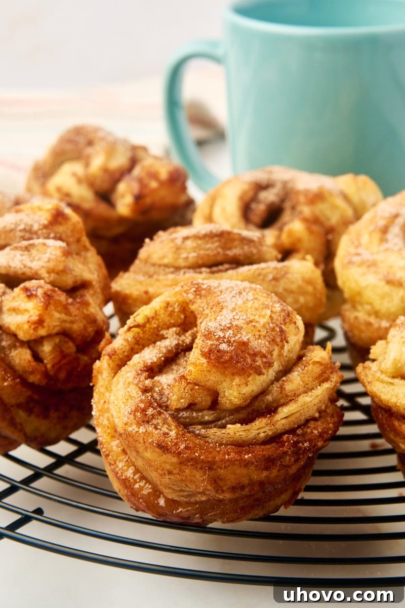 A close-up of a stack of golden-brown cruffins cooling on a wire rack, with a rustic blue coffee mug blurred in the background, suggesting a perfect pairing.