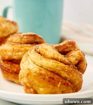 A close-up view of a perfectly baked cruffin on a white plate, with more cruffins and a coffee mug softly blurred in the background.