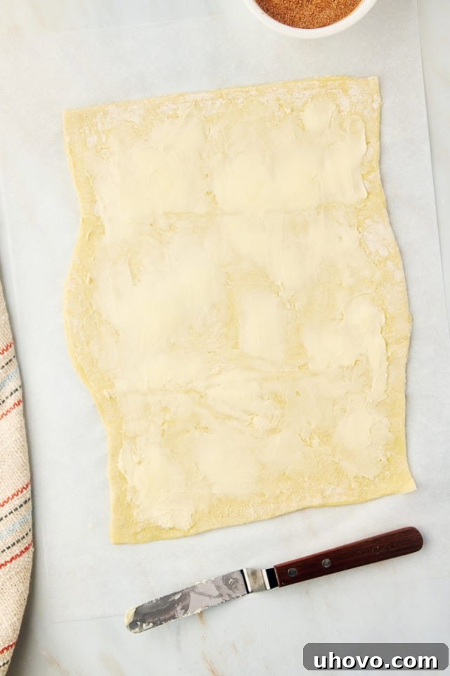 An overhead view showing a sheet of puff pastry expertly rolled out, with a thin, even layer of softened butter being spread across its surface.