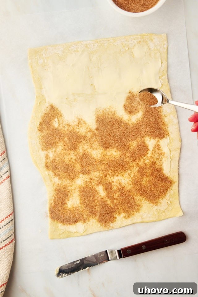 A close-up, overhead shot of the buttered puff pastry being generously sprinkled with the cinnamon-sugar mixture, ensuring even coverage.