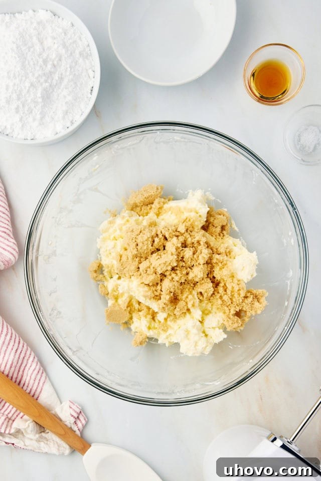 Overhead view of brown sugar being added to the creamed butter and cream cheese mixture in a glass mixing bowl.