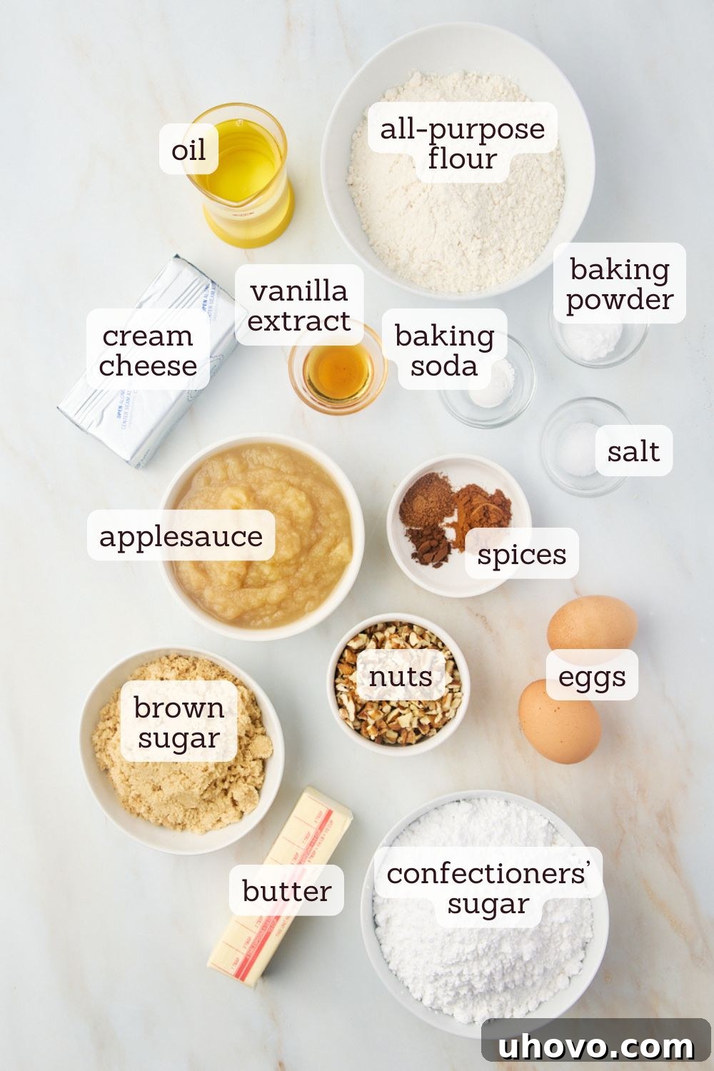 Overhead view of labeled ingredients laid out for making applesauce cake, including flour, spices, oil, brown sugar, eggs, applesauce, and vanilla extract.
