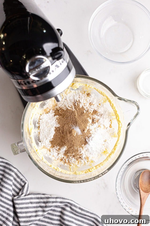 Overhead view of confectioners' sugar, heavy cream, chai spice blend, vanilla extract, and salt added to the beaten butter in a stand mixer bowl.