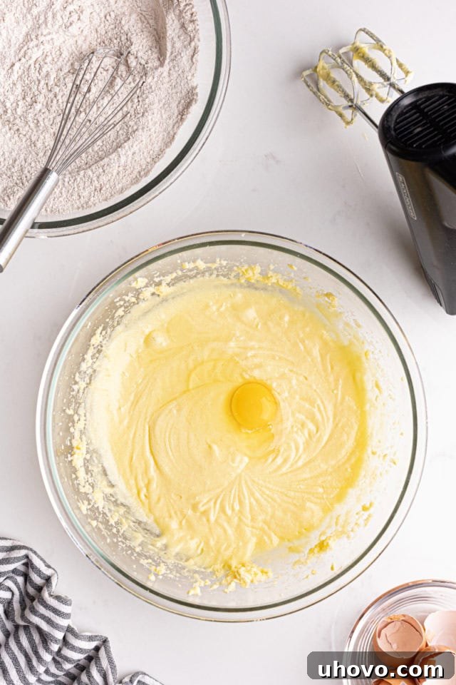 Overhead view of an egg being added to the creamed butter and sugar mixture in a mixing bowl.