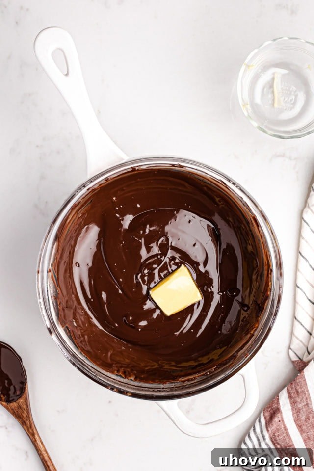 Melted chocolate in a bowl, with a piece of unsalted butter being stirred in to achieve a smooth and glossy finish for the topping.