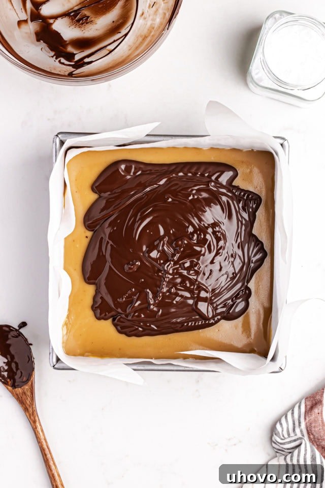 Smooth, melted chocolate being spread evenly over the firm caramel layer in the square baking pan, forming the final top layer of the Millionaire Shortbread Bars.