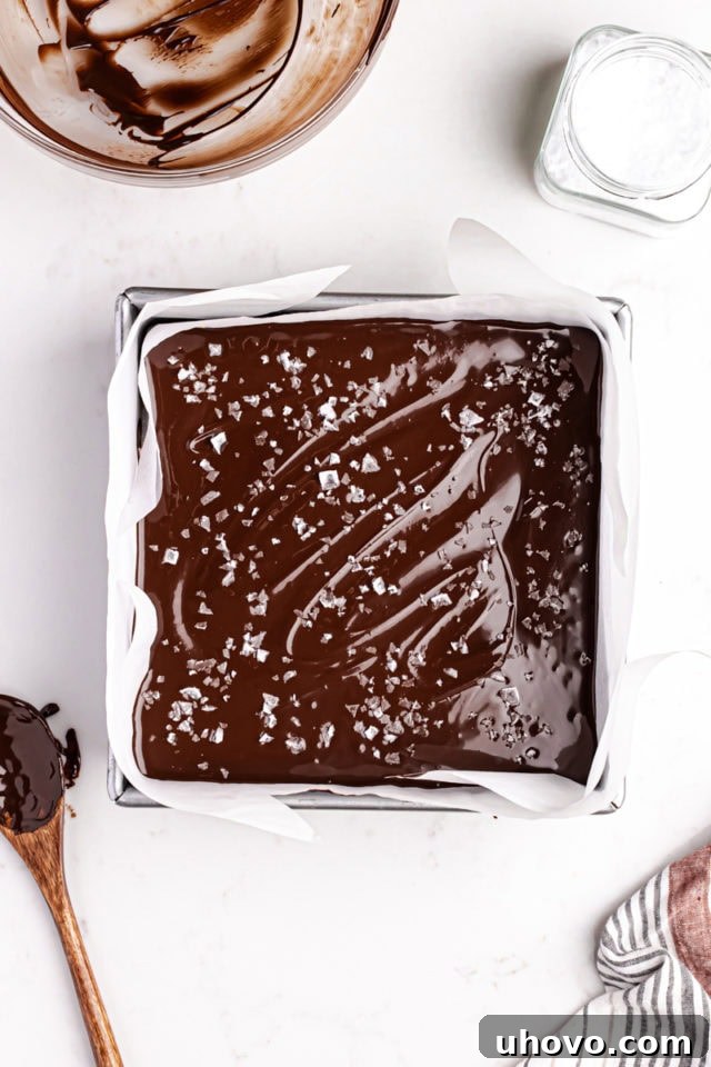 The top chocolate layer of Millionaire Shortbread Bars, beautifully covered with a delicate sprinkle of flaky sea salt, adding a gourmet touch before chilling.