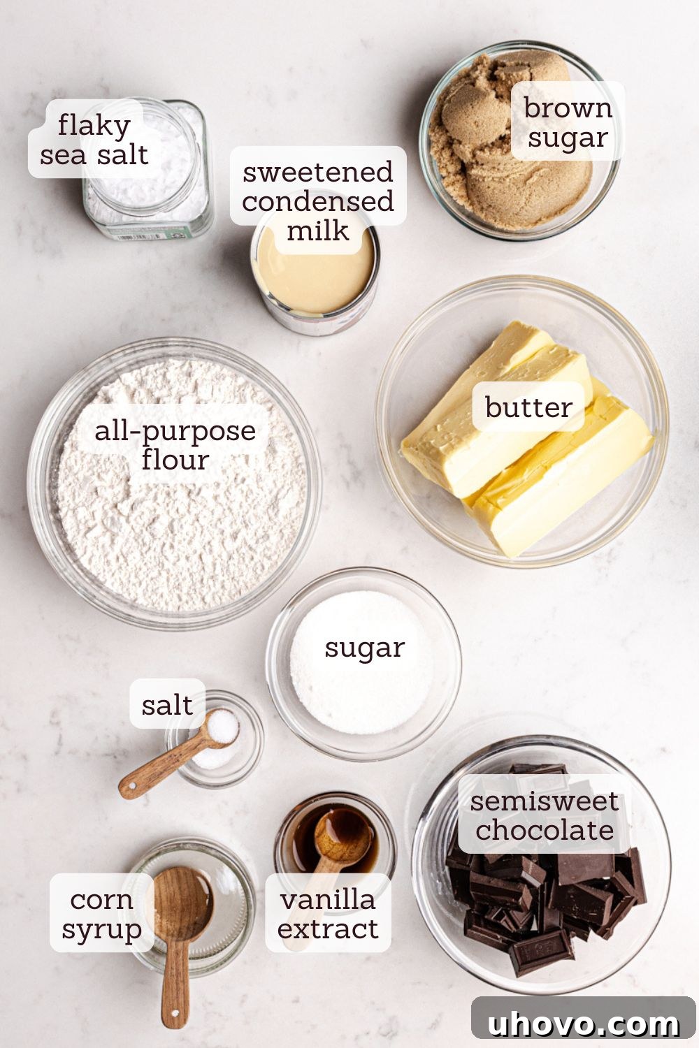 Overhead view of various ingredients for Millionaire Shortbread Bars, neatly labeled and arranged on a kitchen counter, including butter, sugar, flour, condensed milk, and chocolate, ready for baking.