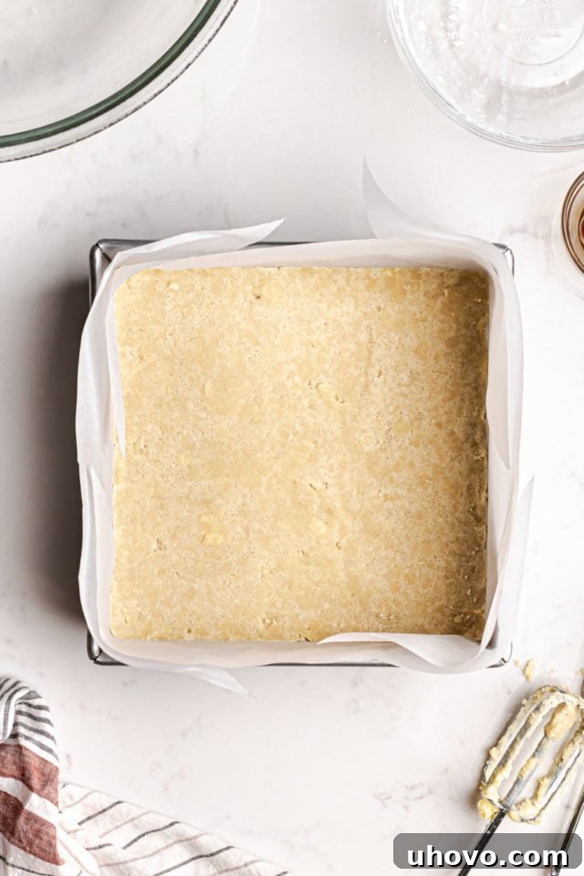 The shortbread dough being pressed firmly and evenly into an 8-inch square baking pan lined with parchment paper, ensuring a compact and smooth base.