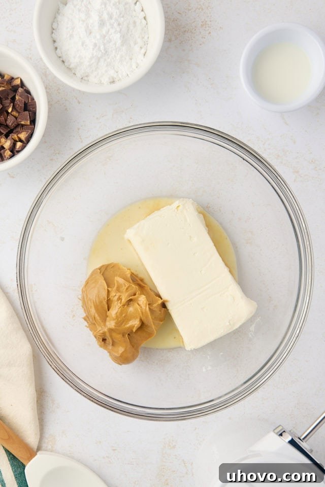 Overhead view of soft cream cheese, creamy peanut butter, and a splash of milk being beaten together in a clear glass mixing bowl, forming a smooth base for the dip.