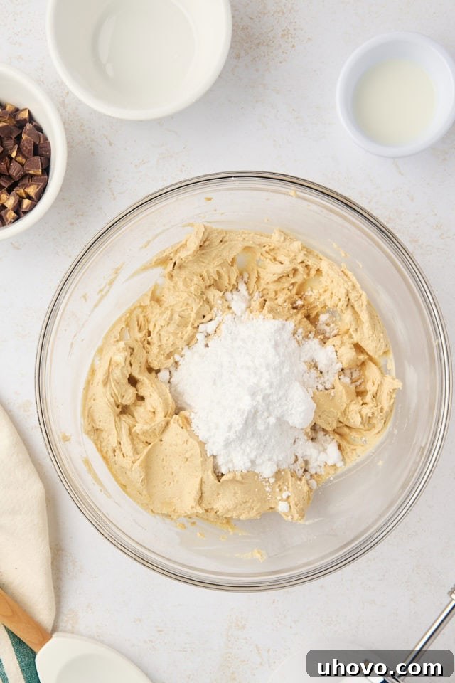 A generous amount of white confectioners' sugar being gradually added to the smooth cream cheese and peanut butter mixture in a glass bowl, ready to be incorporated.