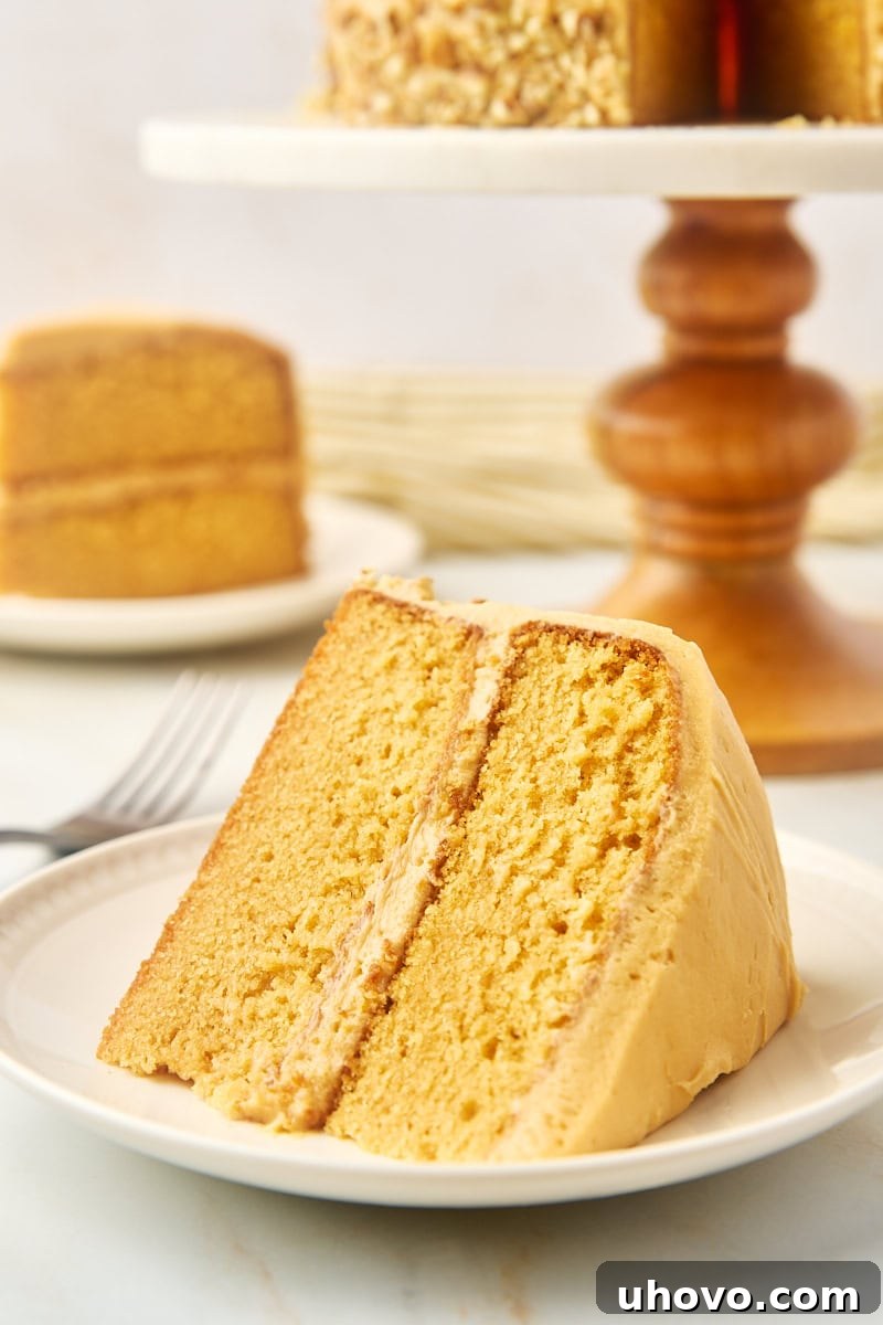 A beautifully decorated slice of dulce de leche cake on a white plate, with a larger frosted cake in the soft background, highlighting its rich, caramel-like goodness.