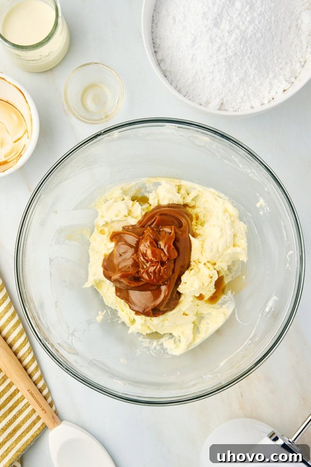 An overhead view of softened butter being beaten in a glass mixing bowl, with dulce de leche and vanilla extract ready to be added for the frosting base.