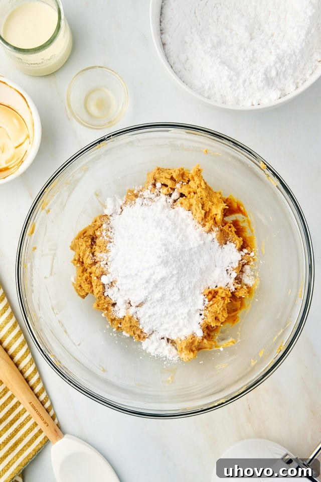 An overhead view of confectioners' sugar being gradually added to the dulce de leche frosting mixture in a glass mixing bowl, showing the thickening process.