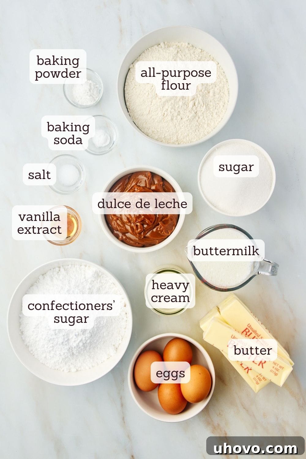 An overhead view of carefully measured ingredients for making dulce de leche cake laid out on a clean surface, including flour, butter, eggs, sugar, and a jar of dulce de leche.