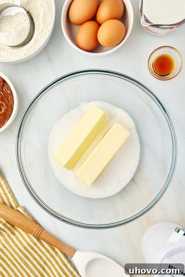 A close-up, overhead view of softened butter and granulated sugar being creamed together in a clear glass mixing bowl, showcasing the light, fluffy texture being developed.