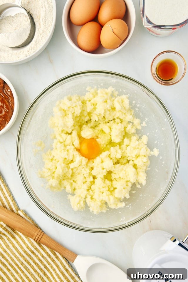 An egg being carefully added to the creamed butter and sugar mixture in a glass mixing bowl, illustrating the step of incorporating eggs one at a time for proper emulsification.