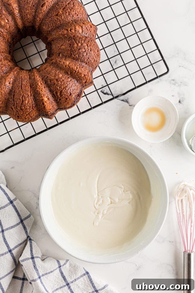 A small bowl of creamy, white glaze placed next to a freshly baked cinnamon Bundt cake, still warm from the oven.