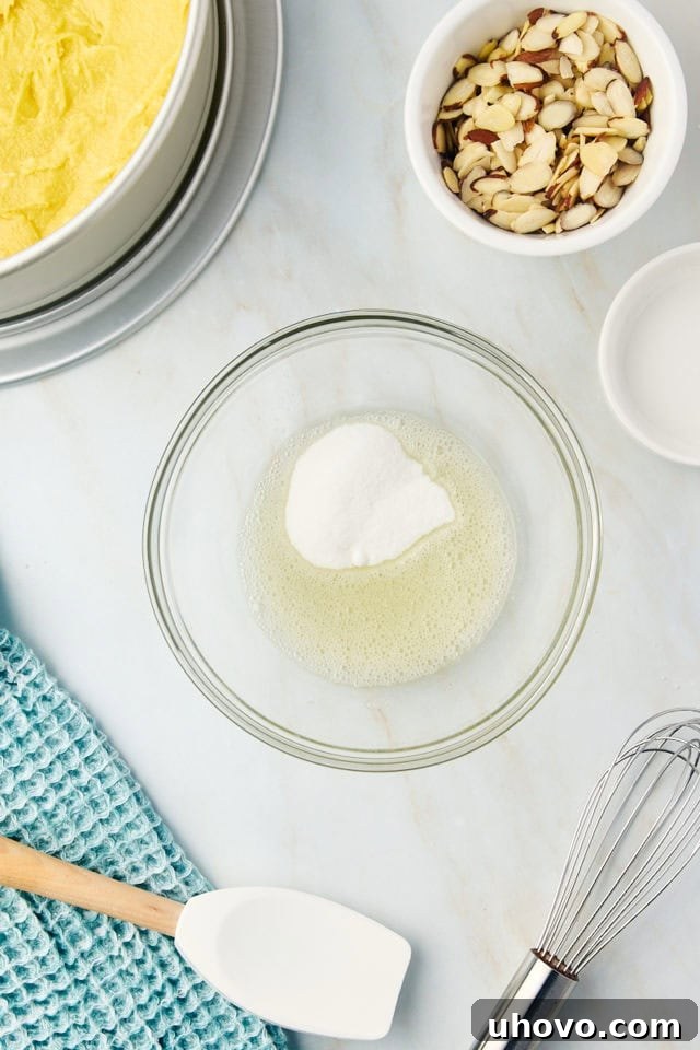 Overhead view of granulated sugar being stirred into the frothy egg white in a small glass bowl, creating a glossy mixture for the almond topping.