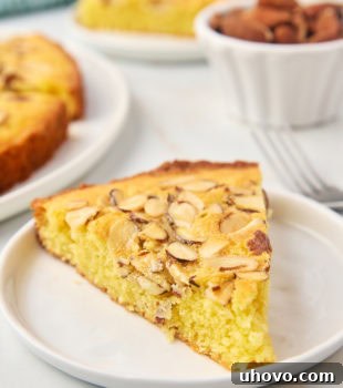 A slice of almond torte on a white plate with more cake in the background, showing the golden crunchy topping.