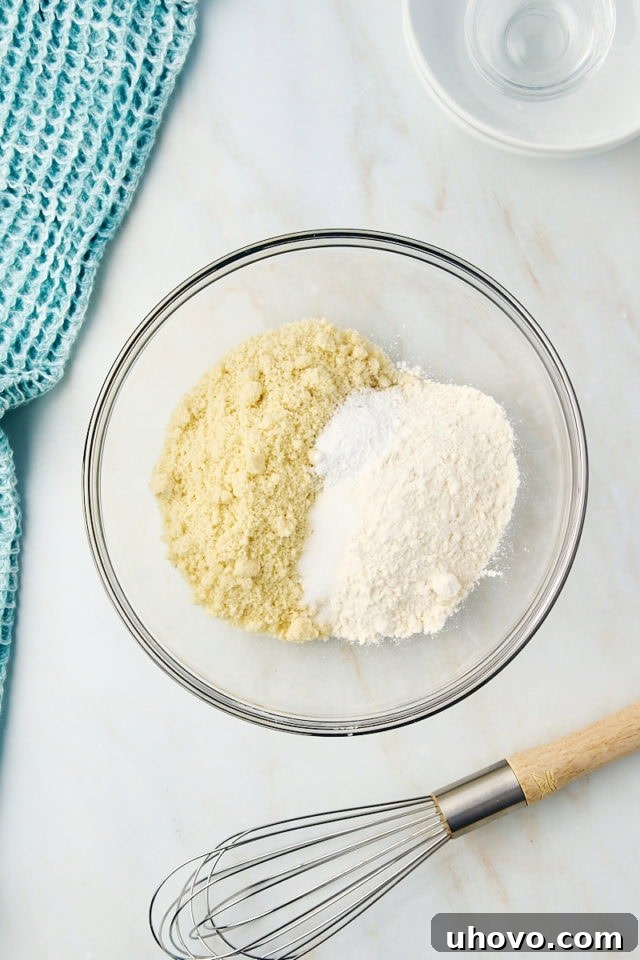 Overhead view of almond flour, all-purpose flour, salt, and baking powder perfectly measured and combined in a clear glass mixing bowl, ready for the next step.