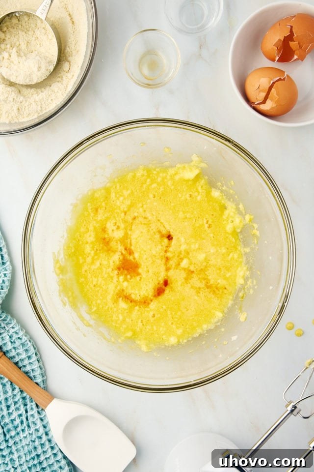 Overhead view of almond and vanilla extracts being poured into the moist cake batter, showing the clear liquids blending into the yellow mixture.