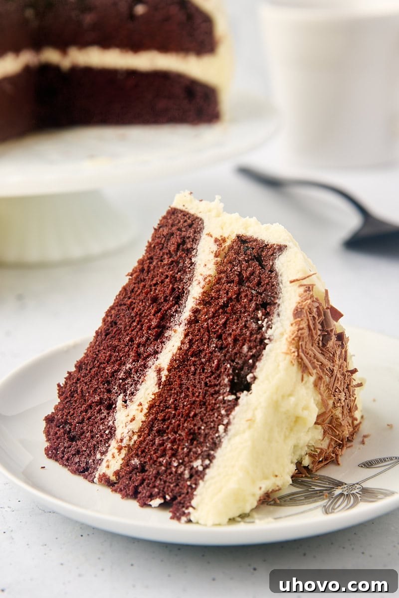 A perfectly sliced piece of mahogany cake on a white plate, with the rest of the cake blurred in the background, showcasing its moist crumb and creamy ermine frosting.