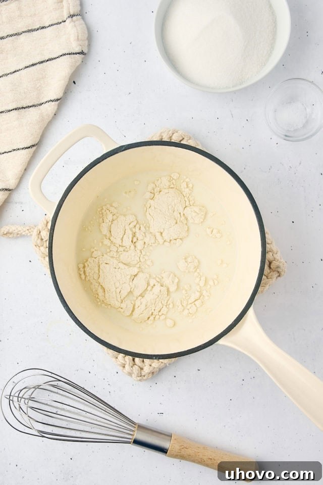 Overhead view of milk and all-purpose flour being whisked together in a small saucepan over heat, beginning the ermine frosting process.