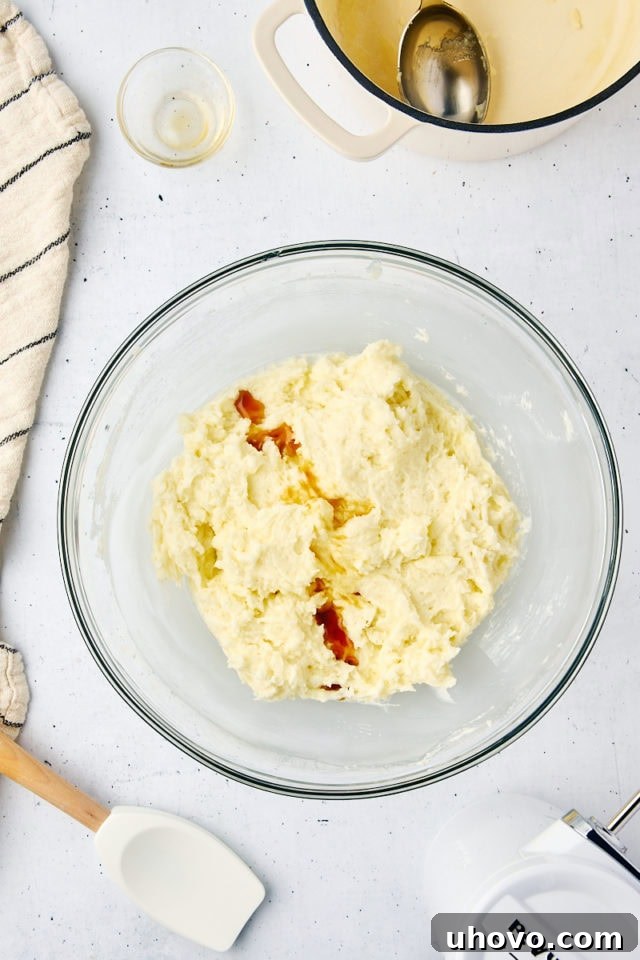 Overhead view of vanilla extract being streamed into the developing ermine frosting in a mixing bowl, for final flavoring.