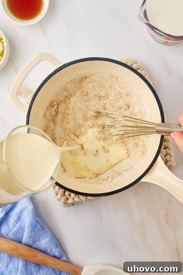 An overhead shot showing heavy cream being slowly poured into the sugar and cornstarch mixture in a saucepan, being whisked gently.