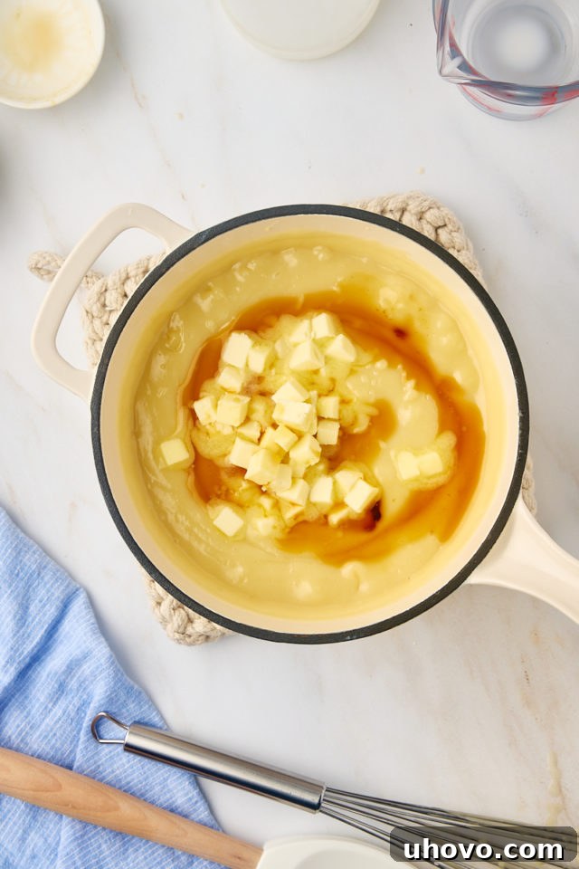 Butter cubes and vanilla extract being stirred into the hot, thickened sugar cream pie filling in a saucepan.