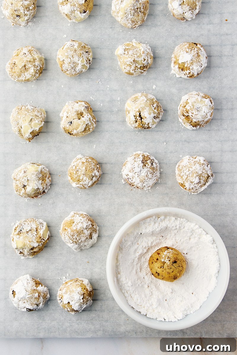 Close-up overhead view of warm snowball cookies being generously rolled in a bowl of fresh powdered sugar, creating their iconic snowy coating.