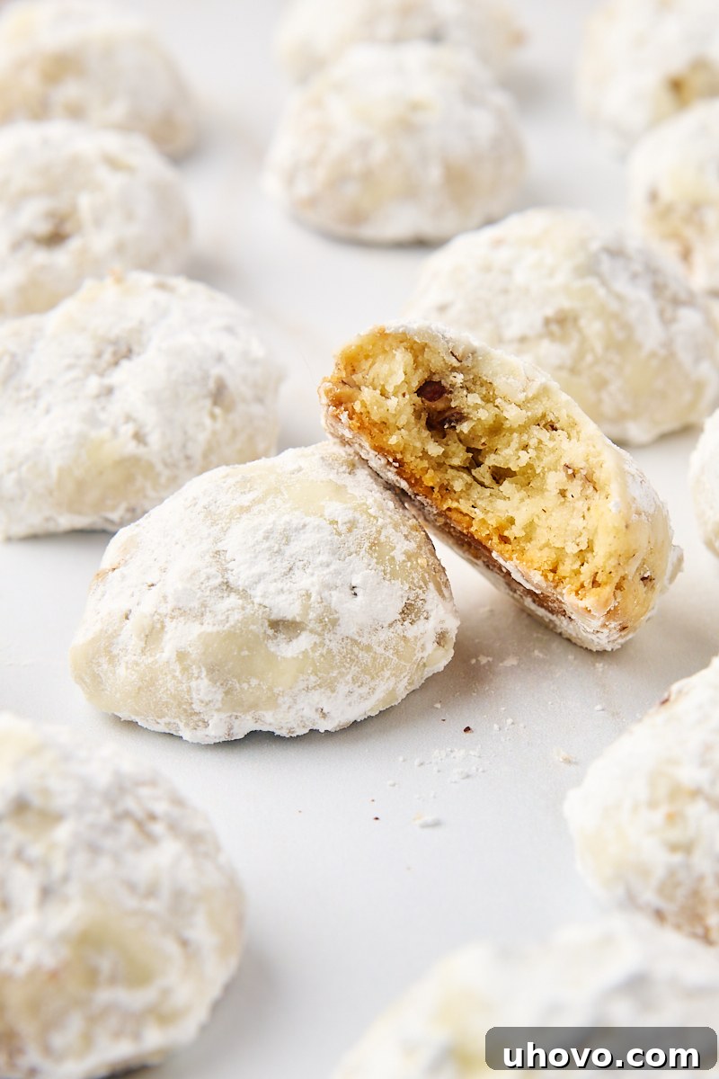 A beautifully composed shot featuring a halved snowball cookie, revealing its tender, nutty interior, propped against another cookie, surrounded by more perfect powdered sugar snowballs.