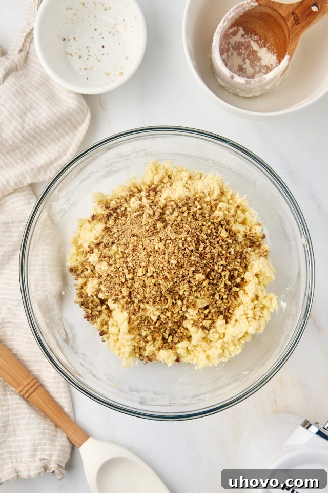 Finely ground toasted nuts being added to the crumbly snowball cookie dough in a mixing bowl.